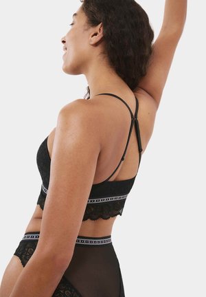 Bralette de encaje negro con un dobladillo con forma de ondas, tirantes ajustables y una banda en contraste que presenta un patrón de logo. Textura suave y lisa.