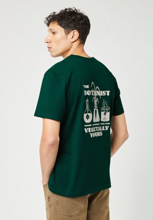 T-shirt en coton vert avec un imprimé dans le dos présentant des illustrations botaniques et du texte en blanc, col rond, manches courtes.