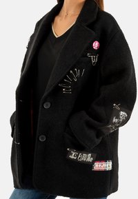 Cappotto oversize in misto lana nero con spille di sicurezza, toppe grafiche e bottoni. Le texture notevoli includono uno strato esterno morbido e peloso.