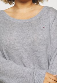 Pull en tricot gris avec un col large et des manches longues. Présente un petit logo brodé en rouge et bleu sur la poitrine. Texture douce.