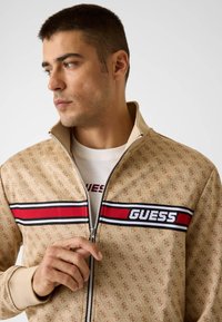 Giacca beige con zip caratterizzata da un motivo con logo ripetuto, strisce rosse e nere a contrasto con testo "GUESS" e polsini a coste.