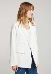Blazer blanc à double boutonnage avec un col cranté, des poches avant et une texture lisse, associé à un haut blanc et un jean bleu.