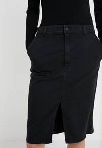 Jupe en denim noire avec une coupe droite, fente à l'avant, deux poches latérales et passants de ceinture. Design classique avec une fermeture à bouton à la taille.