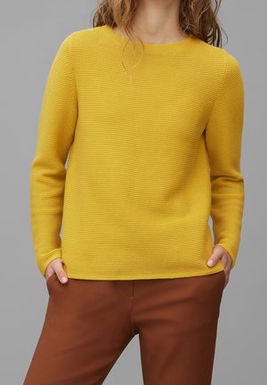 Person trägt einen strukturierten senfgelben Langarmpullover und braune Hose, Hände in den Taschen, vor einem einfarbigen grauen Hintergrund.
