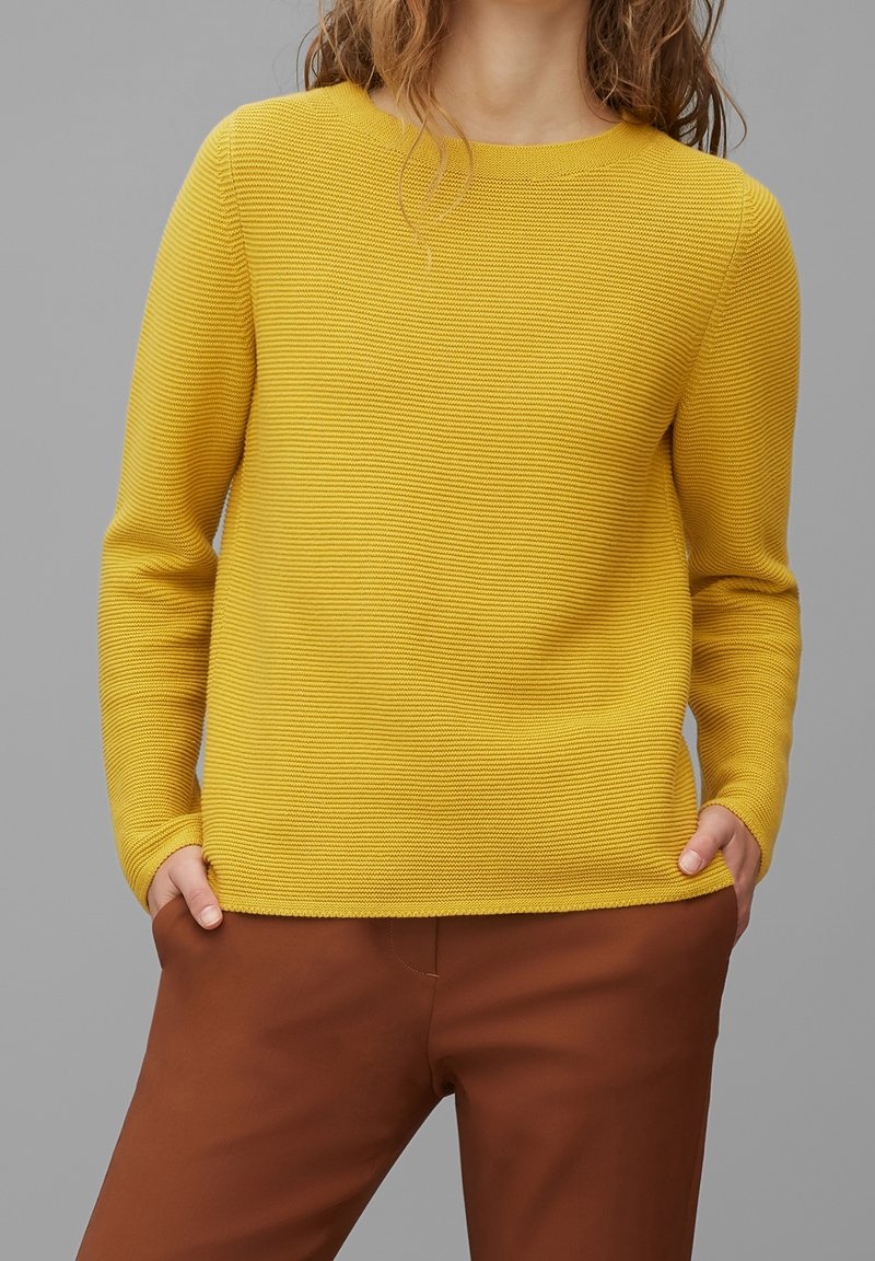 Personne portant un pull à manches longues texturé jaune moutarde et un pantalon marron, mains dans les poches, sur un fond gris uni.