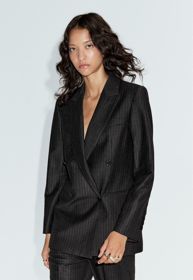 Blazer nero a righe sottili con design doppiopetto, rever affilati e texture liscia. Presenta due tasche laterali e dettagli con bottoni.