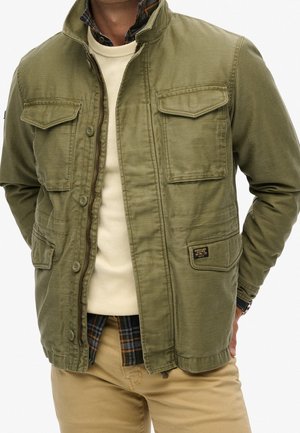 Mann trägt olivgrüne, geknöpfte Jacke über beigem Pullover und beiger Hose mit den Händen hinter dem Rücken vor weißem Hintergrund.