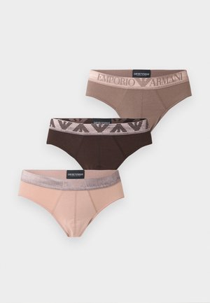 Homme mince à la peau foncée debout pieds nus portant un slip rose clair avec une ceinture gris foncé sur un fond blanc uni.