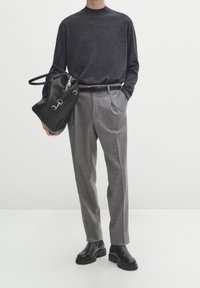 Haut noir à manches longues en tricot, pantalon en laine gris cintré, ceinture noire, tenant un sac en cuir noir, et portant des bottes noires à semelles épaisses.