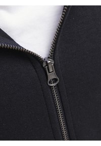 Zwarte sweatshirt met rits en een textuurstof; heeft een metalen rits met een plastic trekker, gedeeltelijk geopend om een wit interieurshirt te onthullen.