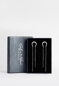 Silberne Creolenohrringe mit Kettenanhänger, präsentiert in einer strukturierten schwarzen Schminkbox mit weißem Logo auf dem Deckel.