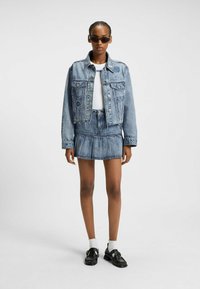 Denim jasje en rokset in lichtblauw, met een geplooid ontwerp, gestructureerde pasvorm en knoopdetails. Gepaard met zwarte schoenen en een zonnebril.