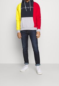 Sudadera con bloques de color que presenta un cuerpo azul marino con mangas rojas y amarillas, una sección inferior gris y un logotipo bordado. Combinada con jeans oscuros y zapatillas blancas.