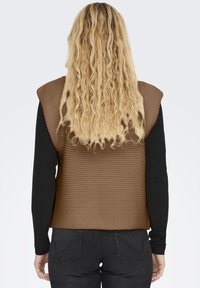 Bruine gebreide vest met een gestructureerd patroon, ronde schouders en een open rug, gedragen over een zwart shirt met lange mouwen, gecombineerd met donkere jeans.