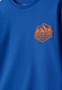 Zila T-krekla ar oranžu emblēmu, uz kuras attēloti kalni, koki, ugunskurs un teksts "PIEDZĪVOJUMS SAVVAĻĀ" uz krūšu daļas.