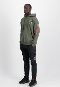Khaki zöld kapucnis pulóver NASA logóval, zsinóros kapucnival és ujjra varrt pachel. Fekete cargo nadrág zsebekkel, slim fit szabással, és fekete sportcipőkkel.