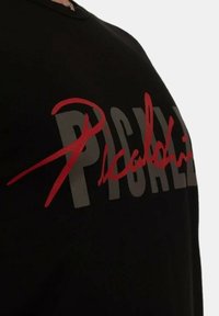 Schwarzes T-Shirt mit großem grauem und rotem Text. Der Text enthält ein auffälliges Unterschriftsdesign, das den Markennamen betont.