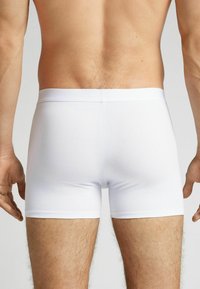 Bílé boxerky z elastického bavlněného materiálu s měkkým elastickým pasem, ukazující přiléhavý design a hladkou texturu z pohledu zezadu.