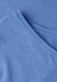 Modrá textilní blůza s texturovaným povrchem, V-výstřihem a hladkými okraji. Barva je jednotná s jemnými variacemi v textuře.