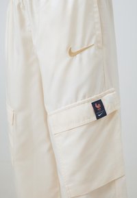 Béžové cargo nohavice s hladkým, lesklým povrchom. Obsahujú zlaté logo Nike a tmavomodrý štítok s emblémom kohúta. Sú vybavené viacerými vreckami.