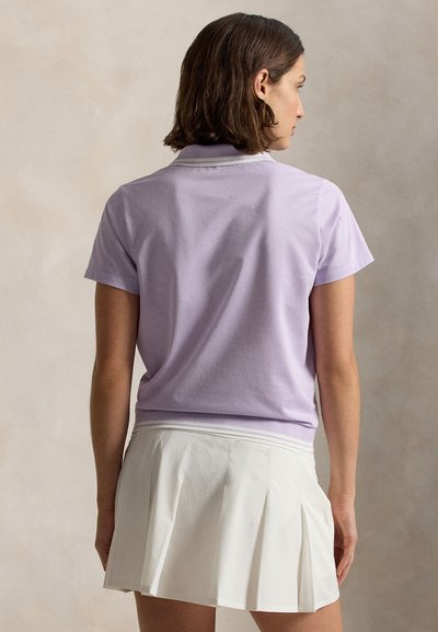 Γυναίκα με κοντά καστανά μαλλιά, ορατή από πίσω, που φορά ανοιχτό μωβ πουκάμισο πόλο με κοντά μανίκια και λευκή πλισέ φούστα.