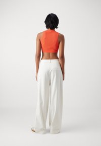 Kobieta stojąca tyłem, ubrana w bezrękawnik w kolorze pomarańczowym oraz białe, wysokie spodnie o szerokich nogawkach z białymi butami, na jednolitym tle.