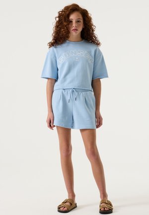 Sudadera de algodón de manga corta en azul claro con la palabra "SUNSET" en letras blancas, combinada con pantalones cortos a juego. Cuenta con cintura ajustable con cordón y un corte relajado.