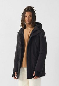 Jongeman met schouderlange dreads, gekleed in een zwarte gewatteerde jas, een beige sweater en witte broek, tegen een eenvoudige lichte achtergrond.