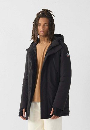 Jongeman met schouderlange dreads, gekleed in een zwarte gewatteerde jas, een beige sweater en witte broek, tegen een eenvoudige lichte achtergrond.