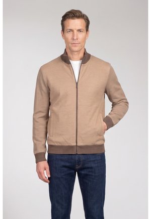 Man draagt beige jas met rits, bruine manchetten en kraag, blauwe spijkerbroek, staat met één hand in de zak tegen een effen achtergrond.
