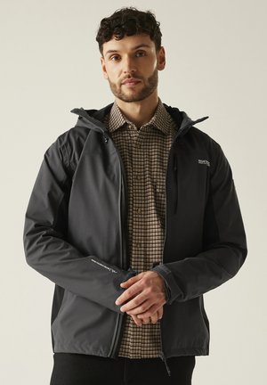 Hombre con chaqueta negra con capucha sobre camisa marrón a cuadros, de pie con las manos entrelazadas al frente sobre un fondo liso.