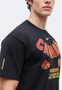 Homme portant un T-shirt noir avec le texte "CANADA" en rouge et jaune, un graphisme de ballon de football avec une feuille d'érable, le nom de la marque NOCTA, et un petit logo Nike jaune.