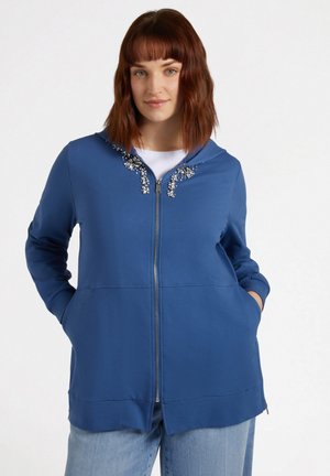 Sudadera azul con cremallera y capucha, que presenta adornos decorativos en el pecho, mangas largas y bolsillos laterales en ángulo. Textura de tela suave.
