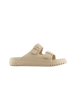 Beige schuiflippers met twee verstelbare gespsluitingen, een dikke zool en een gestructureerde tread voor grip. Gemaakt van zacht, duurzaam materiaal.