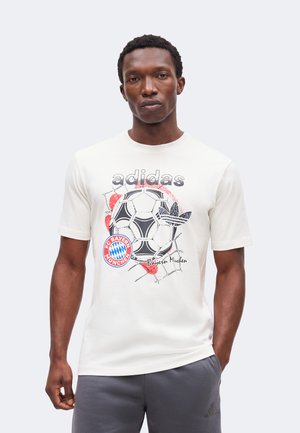 Άνδρας με λευκό t-shirt Adidas με γραφικό μαύρης και κόκκινης μπάλας ποδοσφαίρου και λογότυπο της FC Bayern München, συνδυασμένο με γκρι παντελόνι Adidas.