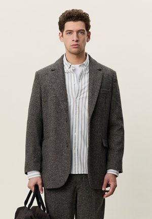 Jeune homme aux cheveux bouclés portant un costume gris sur une chemise rayée, tenant un sac en tissu foncé devant un fond clair uni.