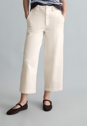 Person, die cremefarbene, weit geschnittene, verkürzte Hosen, dunkelbraune flache Schuhe mit Riemen und ein braun-blaues ärmelloses Stricktop trägt.
