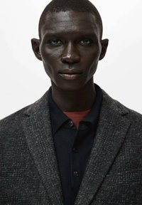 Grauer, strukturiert wirkender Blazer über einem dunklen Poloshirt mit rotem Akzent. Nahaufnahme, mit neutralem Hintergrund, der die Kleidung betont.