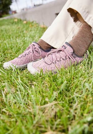Osoba nosiaca béžové nohavice a hnedé ponožky, sediaca na tráve, ukazuje ružové priedušné sieťované tenisky so zaviazanými šnúrkami.