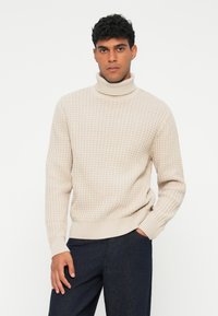 Beżowy sweter z golfem, o fakturowanym wzorze w kratkę, z prążkowanymi mankietami i dołem; noszony z ciemnymi spodniami; model pozujący z jedną ręką w kieszeni.