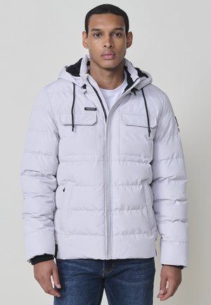 Chaqueta acolchada gris claro con capucha, que presenta cuatro bolsillos frontales, cordones negros y puños acanalados. Detalle de costura notable en toda la prenda.