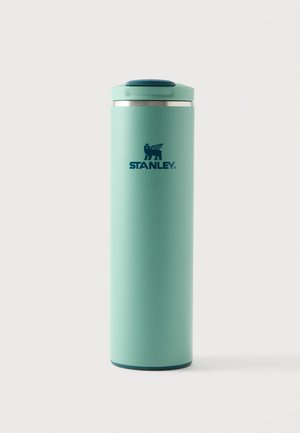 Tazza da viaggio isolata alta di colore verde chiaro del marchio Stanley con coperchio e logo verde scuro su uno sfondo chiaro e semplice.