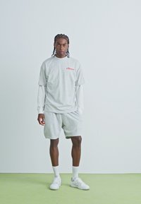 T-shirt en coton gris clair avec un logo rouge, superposé à une chemise à manches longues blanche, associé à un short gris assorti et des baskets blanches.