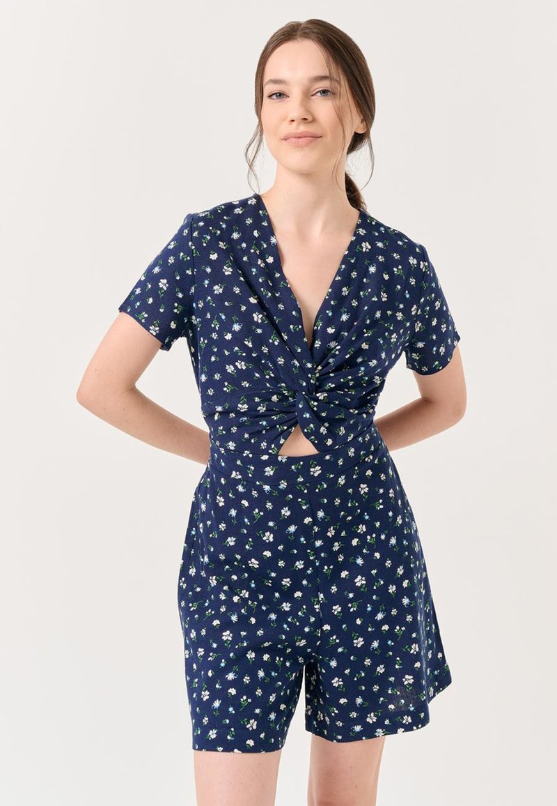 Rompers floreali blu navy con maniche corte, dettaglio a torsione in vita e pantaloni corti. Presenta motivi floreali bianchi e verdi su un tessuto strutturato.