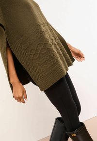 Olivovo zelený pletený pončo s textúrovanými káblovými vzormi, bočnými rozparkami a rebrovaným lemovaním, v kombinácii s čiernymi legínami a lesklými čiernymi čižmami.