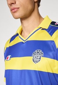 Mand i blå og gul stribede sportsjersey med krave, Champion-logo og NYC CHP 1919 emblem på brystet.