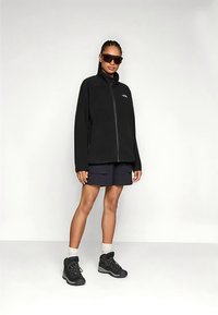 Jachetă din fleece negru cu guler înalt și fermoar frontal, asortată cu pantaloni scurți bleumarin. Modelul poartă bocanci negri de drumeție și ochelari de soare.