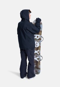 Person in black snowboarding gear and goggles holding a snowboard with forest design and large "ROXY" text, facing sideways on white background.