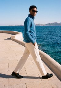Mann mit Sonnenbrille, blauer Jacke, weißen Hosen und schwarzen Schuhen, der an einem klaren Tag einen geschwungenen Steinweg am Meer entlanggeht.