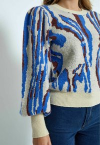 Stickad tröja i krämfärg med blå och bruna abstrakta mönster. Har puffärmar och ribbad nederkant samt mudd. Mjuk textur.